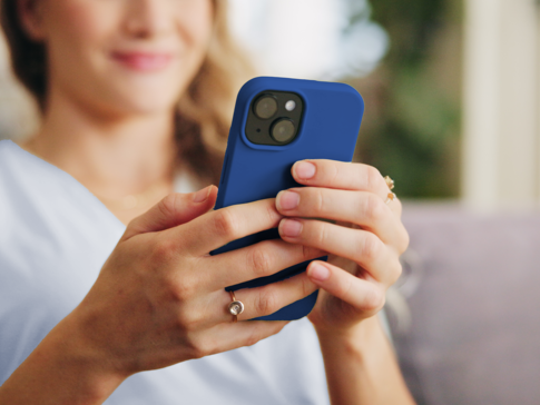 Eine Person hält ein Smartphone mit blauer Schutzhülle in der Hand, während sie auf dem Bildschirm liest oder eine App verwendet. Der Fokus liegt auf den Händen und dem Handy.